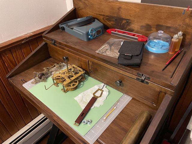 Wooden desk with an open lid exposing two drawers, some clock parts, and a variety of tools
