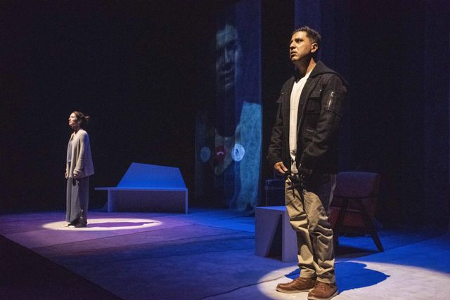 Low side across the stage a white woman and south asian man in spot lights facing the audience -- their faces projected behind them as a face-time call
