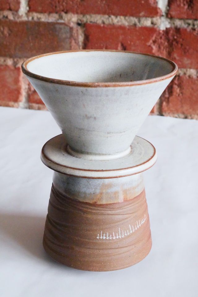 v60 style coffee pour-over with frosty glaze highlighting dark clay that shows through at edges, an inverted cone down to a flat lid resting on the rim of a mug, which is slightly conic the other way - tilting in from a wide base, also in dark clay with the same glaze only at the top. Both have a grooved texture, and there's a row of small white arrows on the mug between grooves