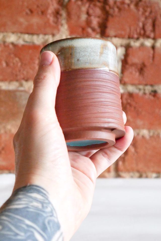 My hand holding up a red-clay cortado-size tumbler with grooved texture, inset foot ring, and white-brown glazed rim. There's just a peak of teal showing inside the foot ring, usually hidden.