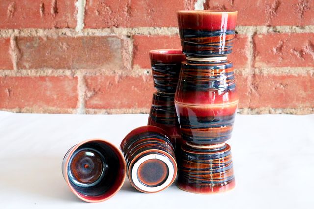 9 tea or espresso cups about 2-3oz, stacked together rim-to-rim, and foot-to-foot - four in front, three behind, and two on the table. A dark dusk glaze breaks red over grooved texture, and has hints of blue between. each has a red rim, and no handle.