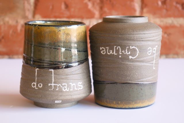Handmade ceramic cups that say 'do trans, be crimes' in a handwritten slab serif. Each has a deep foot ring and slightly rounded wide base, and bare clay textured walls that slope in towards the mossy green-and-gold glazed rim.
