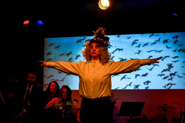 Mrs Reed (Meghan Frank) with arms out wide,
looking stern in front of the band
and a projection of flying bird silhouettes.

