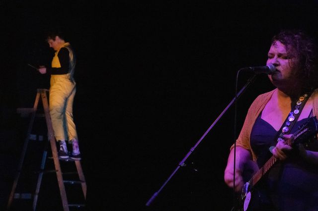 Josie singing in the foreground,
with Miriam on a ladder looking at papers behind her
