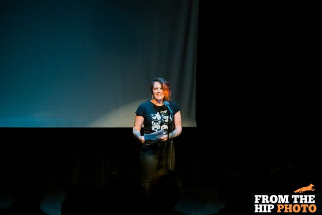 Miriam in a spotlight,
standing at a mic and holding papers,
in front of a large blank screen.
From The Hip Photo watermark logo.
