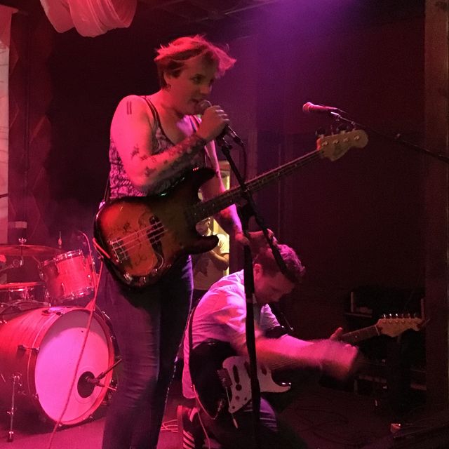 Miriam on stage with a bass under red lights,
singing into a mic,
with Dan on his knees playing guitar,
and a drum set behind them.
