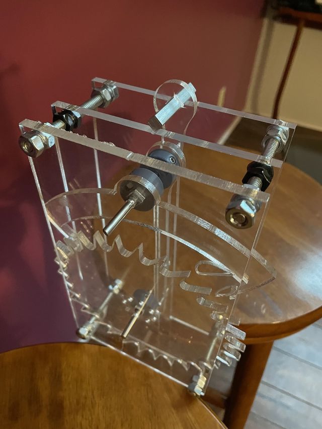 Prototype of a clear acrylic mechanical clock movement, just the pendulum and escape mechanisms, sitting across two stools so the pendulum can hang between